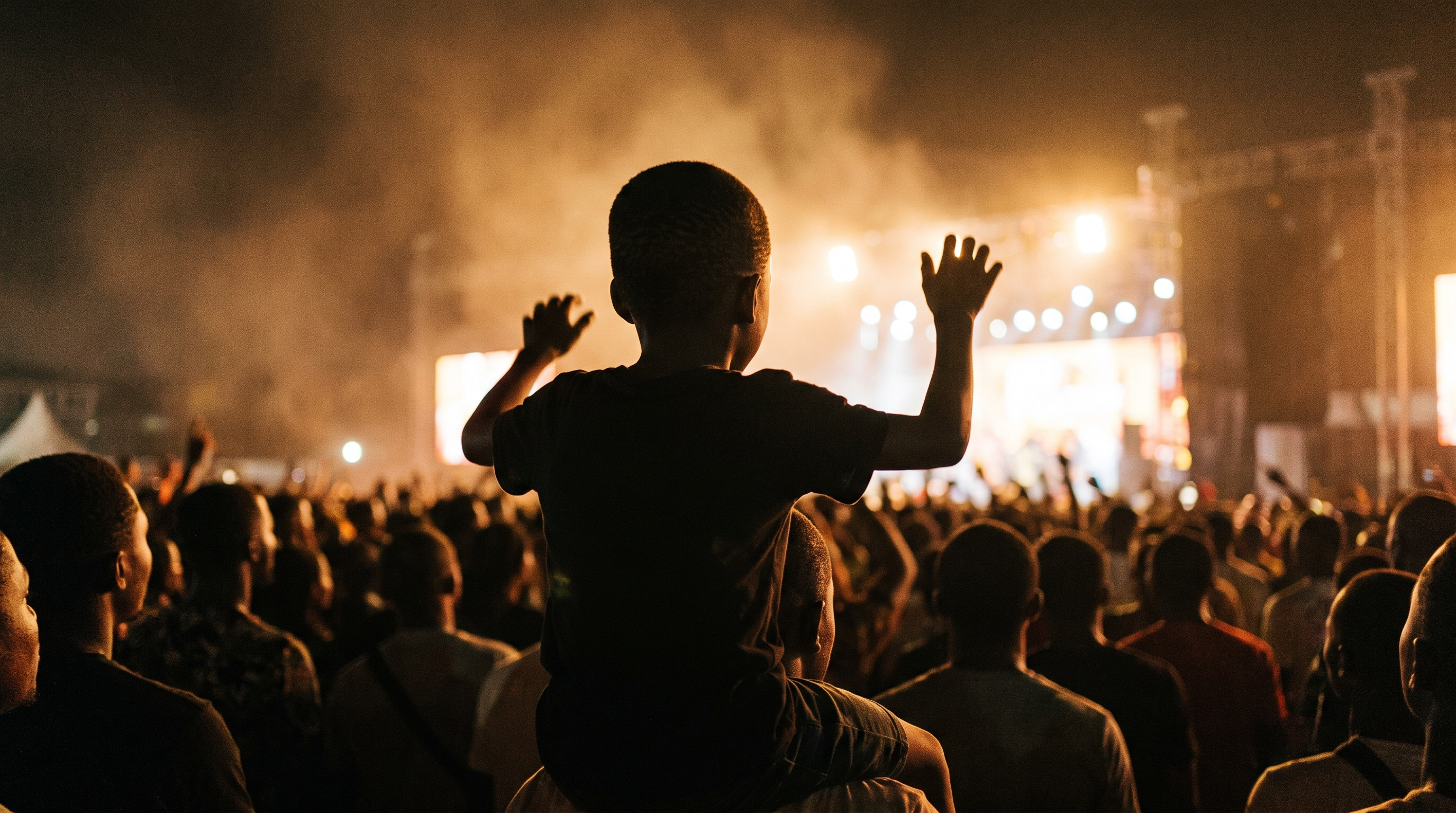 A boy on an adult shoulders in a concert crowd, arms raised towards a lit stage